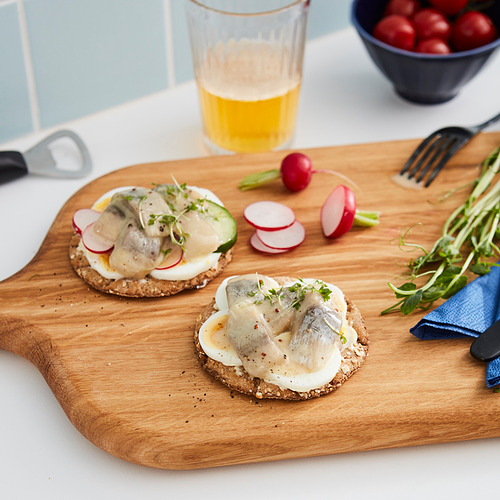La tabla de madera contiene dos sándwiches abiertos cubiertos con arenque, verduras en rodajas y verduras. Vaso de bebida amarilla, cuchillo y rábanos cerca.