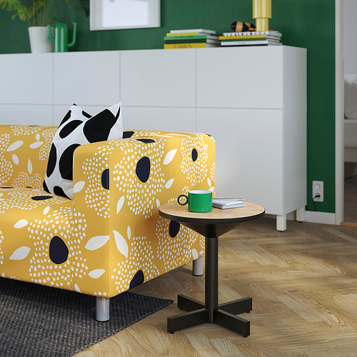 Sofá amarillo estampado con manchas, mesa auxiliar negra, taza verde, libro.