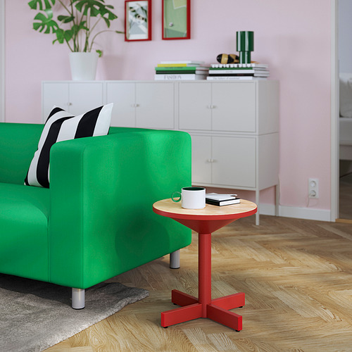 Sofá verde, almohada blanca/negra, mesa auxiliar roja con taza, vasos, libros. Almacenamiento con planta. Paredes rosas, arte. Suelo de madera, alfombra gris.
