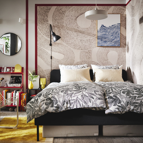Dormitorio moderno con ropa de cama de flores negras, dos almohadas, arte de pared, lámpara, mesa auxiliar, espejo, plantas, libros.