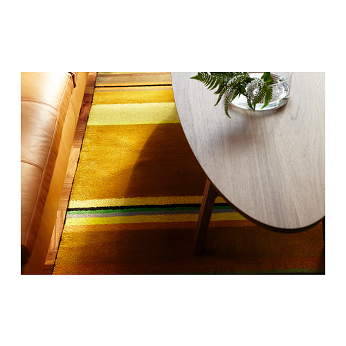 Mesa de centro con tapa de madera y patas metálicas sobre alfombra de rayas, con un jarrón con vegetación.