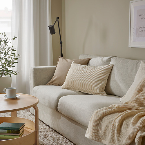 Sala de estar tranquila con sofá ÅSVEIG de color crema, almohadas texturizadas, mesa auxiliar de madera con taza y libros, planta y lámpara de pie.