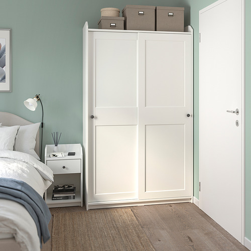 Dormitorio moderno con armario alto HAUGA blanco, cama, mesita de noche, lámpara y libros. Paredes de color azul claro, ambiente relajante.