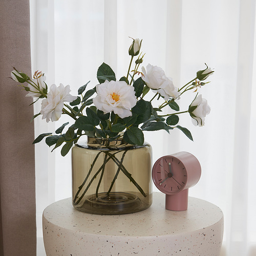 Un jarrón redondo de vidrio con rosas blancas se encuentra en un taburete texturizado junto a un pequeño reloj rosa.