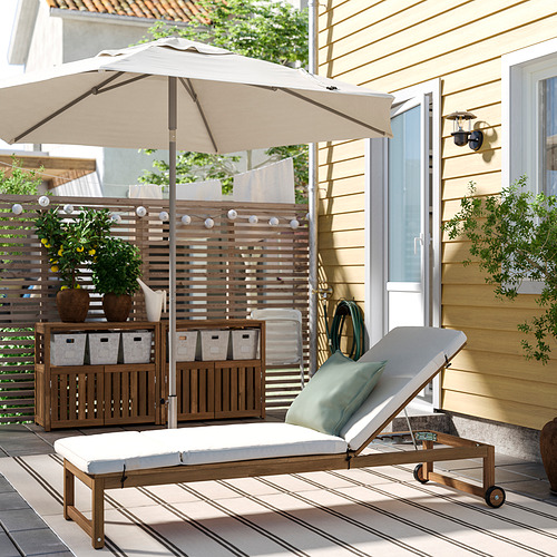 Patio trasero con gran tumbona NÄMMARÖ beige, cojín y almacenamiento de madera, plantas y casa beige.