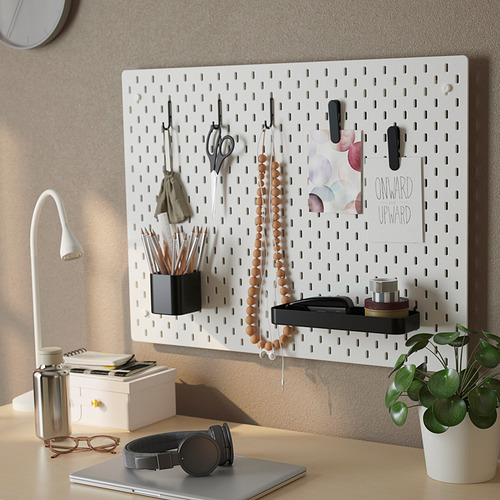 Un organizador de tablero perforado montado en la pared muestra elementos como tijeras, auriculares y una planta con notas, lo que demuestra un almacenamiento eficiente y accesibilidad.
