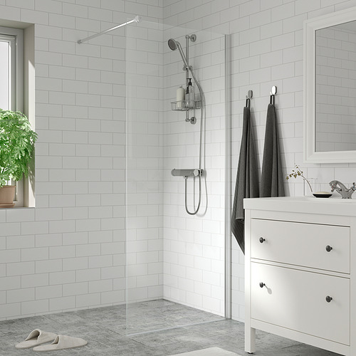 Baño moderno con azulejos blancos, ducha de vidrio, accesorios cromados, toallas grises, lavabo pequeño, espejo y planta.