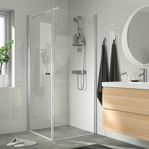 Baño moderno y minimalista con ducha de vidrio, marco plateado delgado, tocador de madera, espejo, toallas negras, plantas, azulejos gris oscuro.