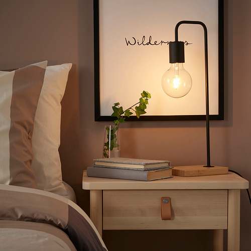 Mesita de noche con lámpara TVÄRHAND, libros y planta. La lámpara es pequeña, negra y moderna. Los libros y las plantas añaden decoración e interés.