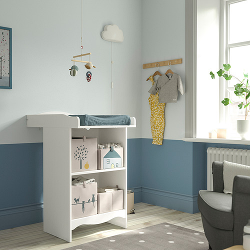 Guardería con cambiador y estantería blancos SMÅGÖRA, decoración azul. La ropa de bebé cuelga por encima, la silla gris a un lado.