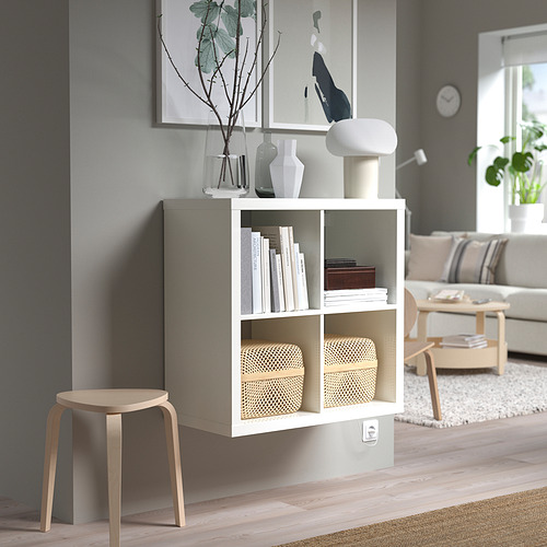 Estantería KALLAX blanca con libros, jarrones, lámpara, junto a taburete, bajo marco de cuadro en pared gris.