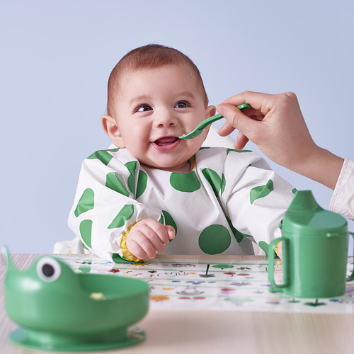 El bebé con babero de manga verde come de cuchara verde, usando un tazón de dinosaurio verde, con alimentación para adultos.
