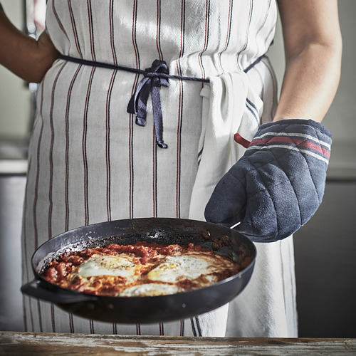 Una persona con un delantal de rayas grises sostiene una sartén de shakshuka con un guante de cocina negro.