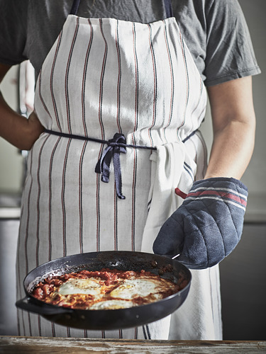 Persona con un delantal a rayas y guantes de cocina que sostiene una sartén con alimentos cocinados.