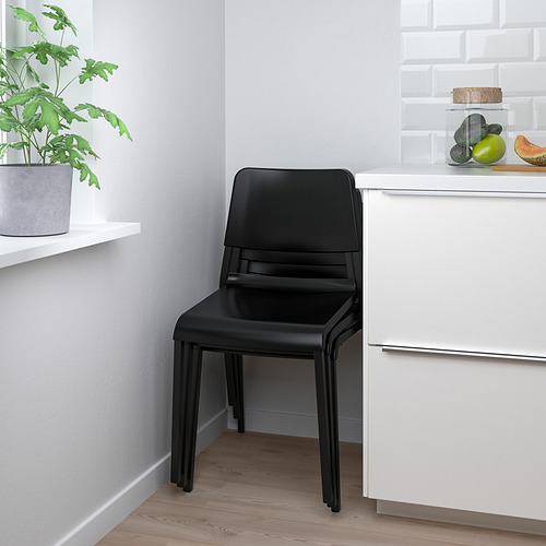 Silla apilable negra, hasta 6 alturas. Adecuado para comedores, entradas o dormitorios. Cerca de cajones blancos y planta.