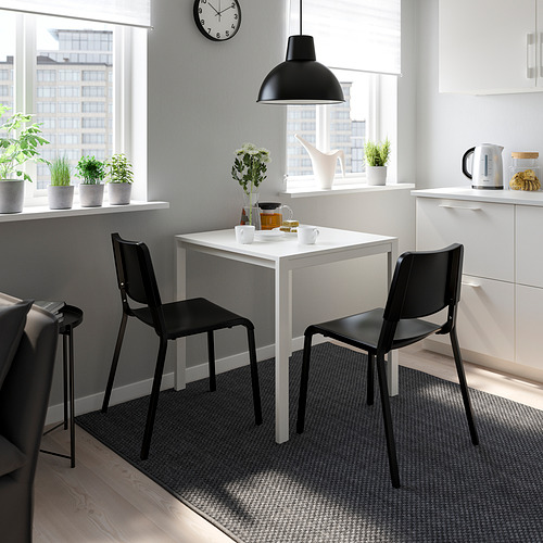 Cocina minimalista con mesa blanca, sillas TEODORES negras, lámpara colgante, plantas y armarios blancos. Las sillas son ligeras y apilables.