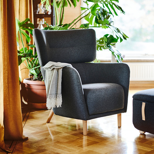 Acogedora silla OSKARSHAMN gris, diseño moderno, patas de madera, cojines de felpa, gran planta en maceta al lado.