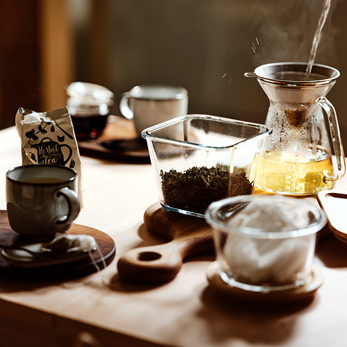 Recipiente de vidrio para alimentos con té suelto e infusor, colocado sobre la mesa con tazas.