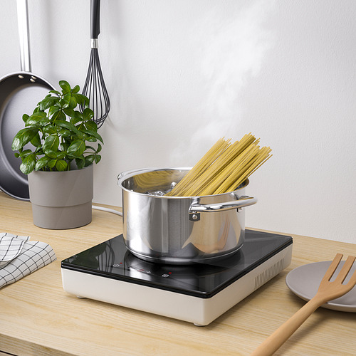 Pequeña configuración de cocina con placa de inducción portátil TILLREDA, cocinando espaguetis. Incluye panel de control, enchufe y decoración de plantas.