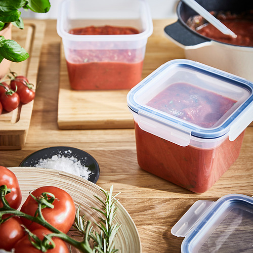 Los recipientes de plástico rojo contienen sopa de tomate, uno vertido en otro, con tomates y hierbas cerca.