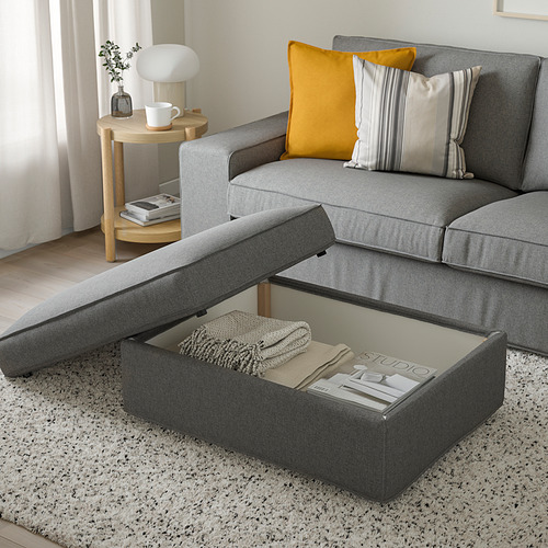 Salón moderno: sofá modular gris con almohada amarilla, mesita auxiliar de madera con lámpara y libros. Otomana abierta, mostrando una manta y revistas en su interior.