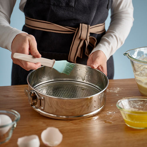 La persona usa un cepillo verde para engrasar un molde desmontable de acero inoxidable, rodeado de ingredientes para hornear.