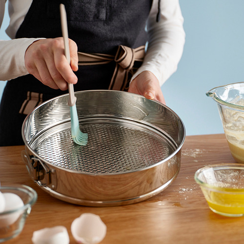 Una persona usando un molde desmontable, cubriéndolo con aceite para hornear. La sartén está sobre una mesa rodeada de ingredientes.
