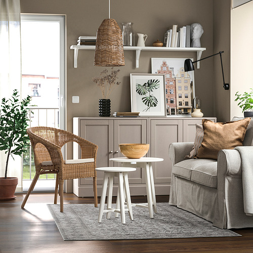 Modern living room with grey sofa, white tables, wicker chair, and wooden decor. Cosy, well-lit, neutral colours.