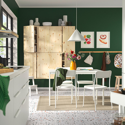 Cocina moderna con paredes verdes, gabinetes de madera clara y sillas blancas apilables alrededor de una mesa. Una alfombra colorida y arte realzan la decoración.