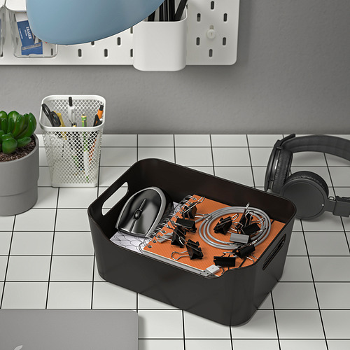 Caja organizadora negra sobre un escritorio con azulejos con auriculares, un mouse y bolígrafos. Ayuda a almacenar y organizar artículos.