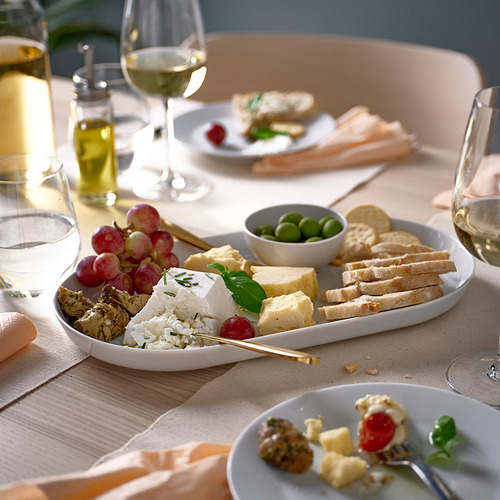 Tabla de quesos en un gran plato blanco con platos más pequeños y copas de vino, rodeados de uvas, aceitunas y pan. Elegante puesta de mesa.