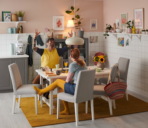 Dos personas cenando en una mesa moderna con sillas BERGMUND amarillas y grises, rodeadas de plantas, cuadros y una lámpara colgante única.