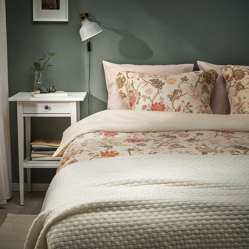 Dormitorio con paredes verdes, ropa de cama blanca, almohadas florales. Pequeña mesita de noche blanca con lámpara, libros y jarrón. Configuración acogedora y ordenada.