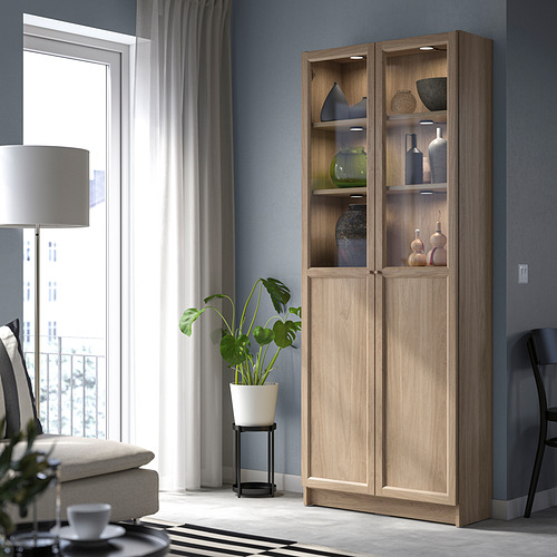 Librería de madera BILLY con jarrones, junto a ventana con sofá y planta.