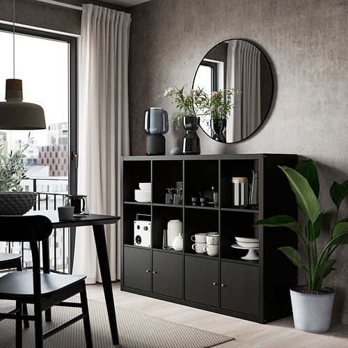 Comedor moderno con estantería KALLAX negra, gran espejo redondo, mesa de comedor negra, sillas y plantas.