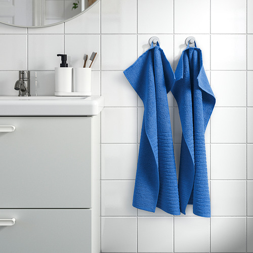 Dos toallas azules VÅGSJÖN cuelgan de la pared del baño, más grandes que los artículos de tocador en la encimera.