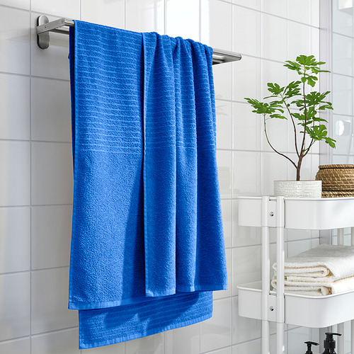 La toalla azul VÅGSJÖN cuelga en el baño, doblada en un riel plateado, más grande que una planta en maceta, estantes blancos cerca.
