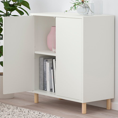 Armario EKET blanco con dos puertas abiertas, que exhibe libros y un jarrón en su interior. Se levanta sobre patas de madera, con una planta en la parte superior.