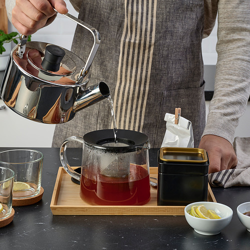 Verter té en una tetera de vidrio con infusor en una bandeja de madera, rodeada de tazas, limones y lata.