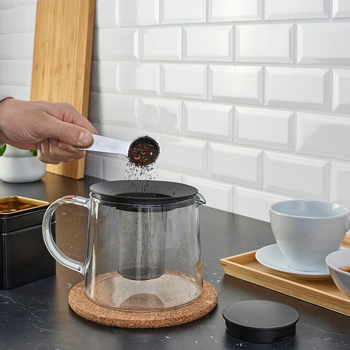 La mano vierte hojas de té en una tetera de vidrio RIKLIG con infusor de malla en el mostrador.