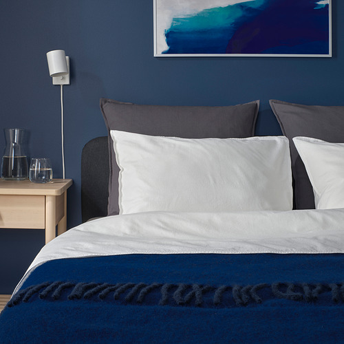 Dormitorio moderno con pared azul oscuro, cojines grises GURLI, ropa de cama blanca, mesita de noche de madera, cristalería azul, lámpara de pared.