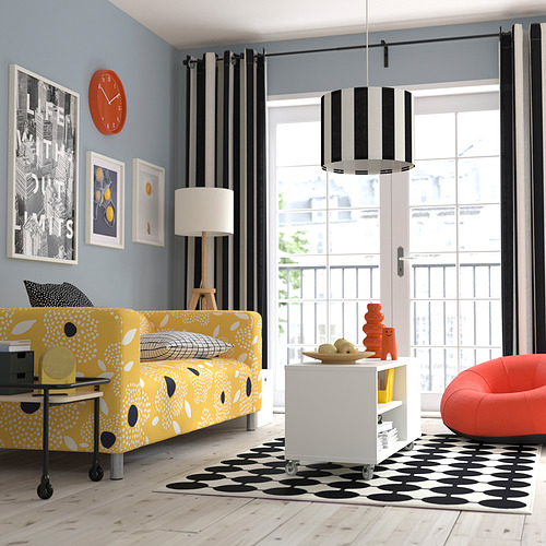 Sofá RINGSTA amarillo, alfombra blanca/negra, reloj naranja, cortinas a rayas. Gran ventana con balcón, lámpara moderna sobre una pequeña mesa con jarrones naranjas.