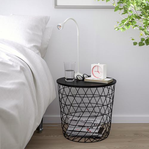 Pequeña mesa auxiliar de alambre negro con patrón de diamante, reloj de sostenimiento, vaso de agua y lámpara de lectura al lado de la cama.
