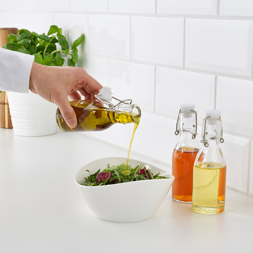 Verter aceite de la botella KORKEN sobre la ensalada. Botellas KORKEN adicionales con líquidos naranjas y amarillos, pared de azulejos blancos.
