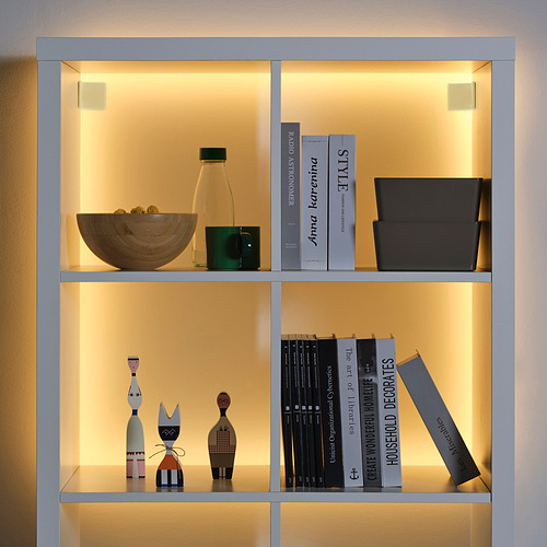 Una estantería iluminada con libros, decoración y tiras de luz KABBLEKA . Los libros se mantienen en pie, la decoración varía en tamaño y la iluminación resalta los objetos.