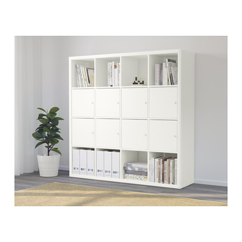 Una estantería blanca KALLAX con libros y armarios, planta al lado, alfombra debajo.