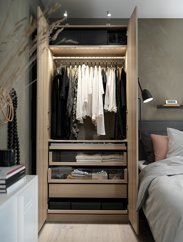 Dormitorio moderno con armario PAX de madera abierto que muestra ropa en blanco y negro, estantes con telas dobladas y cajones de vidrio.