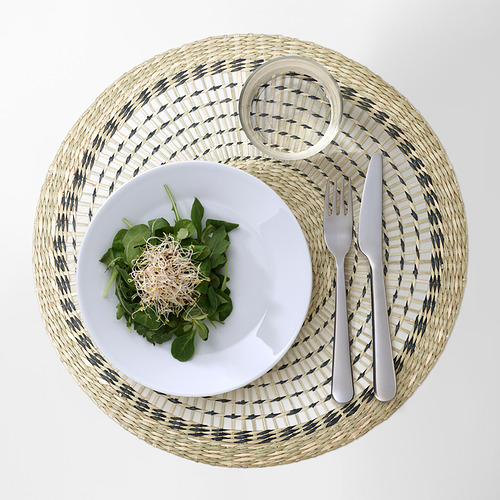 Un mantel individual redondo tejido con un patrón gris y blanco sostiene un plato blanco de ensalada y cubiertos.