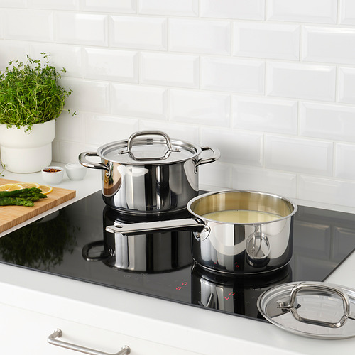 Cocina moderna con cacerolas en placa de inducción, una con tapa, otra con nata. Tabla de cortar con verduras y hierbas, pared de azulejos blancos.
