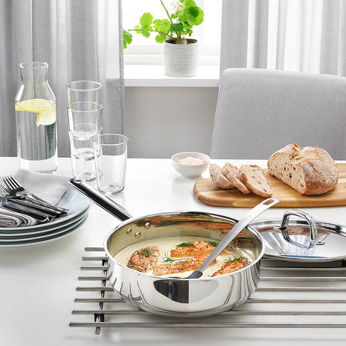 Sartén de acero inoxidable con tapa en la mesa, que contiene alimentos cocinados. Rodeado de platos, vasos y pan, lo que sugiere la preparación de la comida.